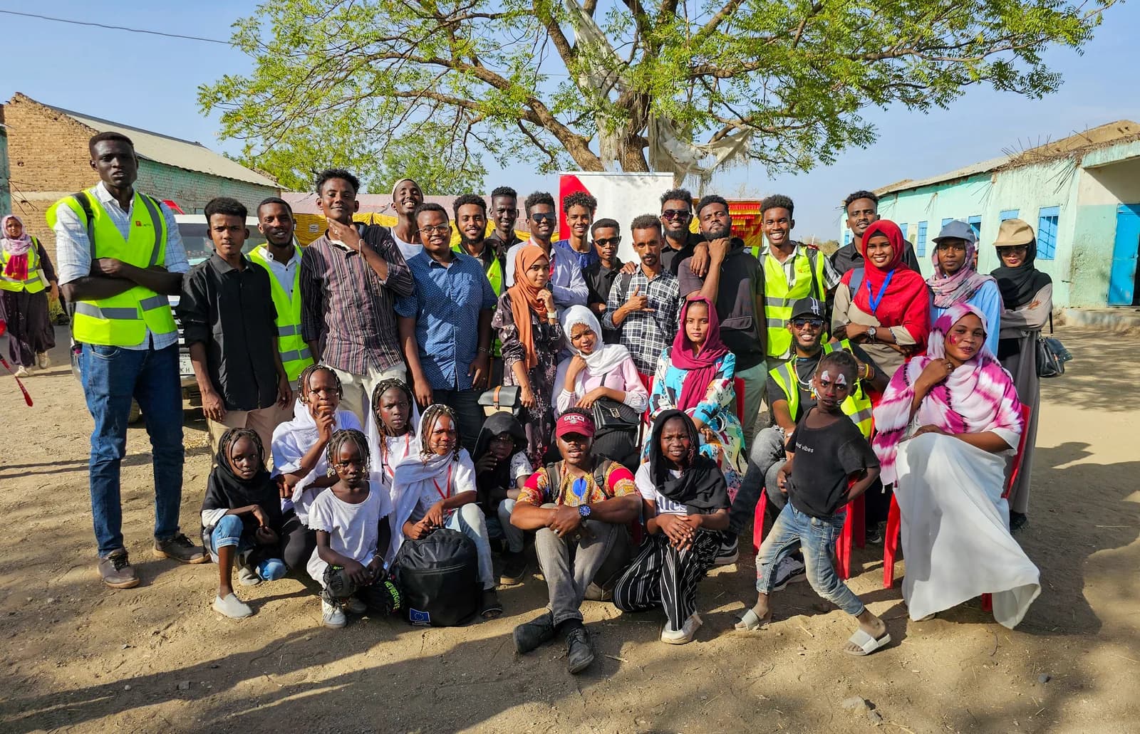 Members of the G.Artistic team and community participants gathered outdoors under a tree, several wearing field vests.